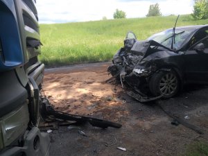 na zdjęciu znajduje się po prawej stronie samochód osobowy koloru ciemnego z wieloma uszkodzeniami a przed nim znajduje się przód samochodu ciężarowego również z uszkodzeniami