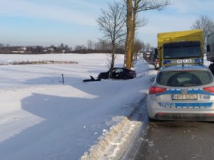 na zdjęciu znajduje się tył oznakowanego radiowozu ustawiony na jezdni gdzie z lewej jego strony w ośnieżonym rowie stoi samochód osobowy przylgnięty drzwiami kierowcy do przydrożnego drzewa,