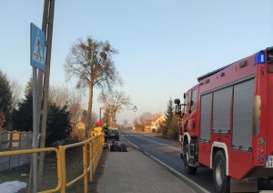 na zdjęciu po prawej stronie na jezdni znajduje się  pojazd Straży Pożarnej a prze nim po lewej stronie na chodniku leży przewrócony motorower, przy którym stoją strażacy