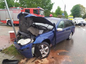 na obrazku znajduje się samochód osobowy ciemnego koloru z uszkodzonym przodem pojazdu, który zatrzymał sie na znaku drogowym