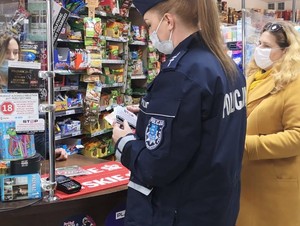 na zdjęciu znajduje się policjantka z naklejką w dłoni, obok niej stoi kobieta, obie stoją przed ladą kasową wewnątrz sklepu monopolowego