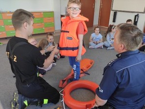 na zdjęciu znajduje się chłopiec, któremu strażak zakłada kapok w kolorze pomarańczowym a obok nich jest policjant z kołem ratunkowym