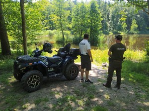 na zdjęciu znajduje się policjant i strażnik leśny stojący w lesie przy akwenie wodnym a obok nich stoi quad