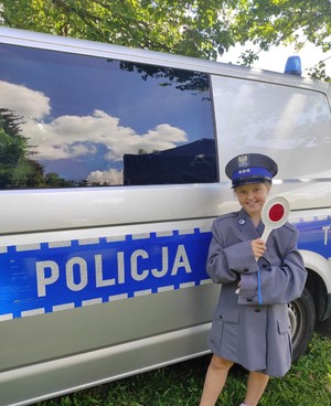na zdjęciu znajduje się dziewczynka ubrana w policyjny mundur galowy z ,,lizakiem&#039;&#039; policyjnym w dłoni na tle radiowozu