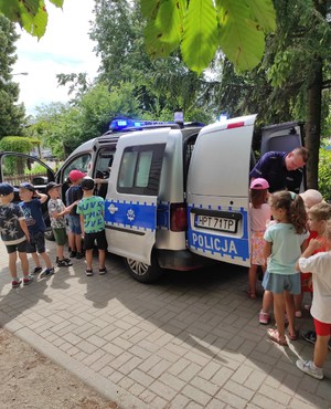 na zdjęciu znajduje się policjant stojący z tyły radiowozu obok którego stoi grupa dzieci