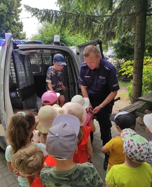 na zdjęciu znajduje się policjant stojący przy policyjnym radiowozie, przed którym stoi grupa dzieci