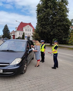 na zdjęciu obok ciemnego samochodu osobowego stoi kobieta wręczająca kierowcy jednorazowy alkotest , nieopodal stoją dwie policjantki