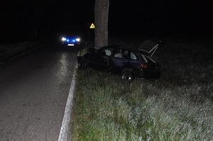 na zdjęciu znajduje się samochód osobowy przy drzewie, przed nim na ulicy stoi radiowóz z włączonymi światłami odblaskowymi, teren zaciemniony nocą