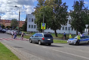 na zdjęciu znajduje się radiowóz policyjny obok którego stoją policjanci, przed nimi znajduje się ulica z przejściem dla pieszych po którym idą osoby, przed przejściem stoi samochód