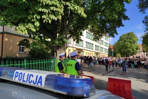 na zdjęciu znajduje się radiowóz policyjny przed którym tyłem stoi dwóch policjantów, przed nimi znajduje się budynek szkoły przed którym stoi grupa osób