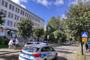 na zdjęciu znajduje się radiowóz policyjny stojący przed przejściem dla pieszych, przed którym stoją osoby, po lewej stronie znajduje się budynek szkoły przed którym na placu stoją dzieci