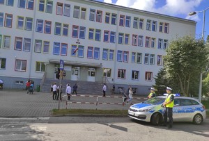 na zdjęciu znajduje się budynek szkolny przed którym na ulicy przed przejściem dla pieszych znajduje się radiowóz obok którego stoją policjanci