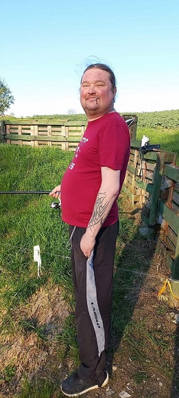 na zdjęciu znajduje się mężczyzna stojący bokiem na trawie ubrany w czerwoną koszulkę i ciemne spodnie dresowe, na lewym przedramieniu widnieje tatuaż, mężczyzna ma związane włosy do tyłu, na brodzie ma lekki zarost
